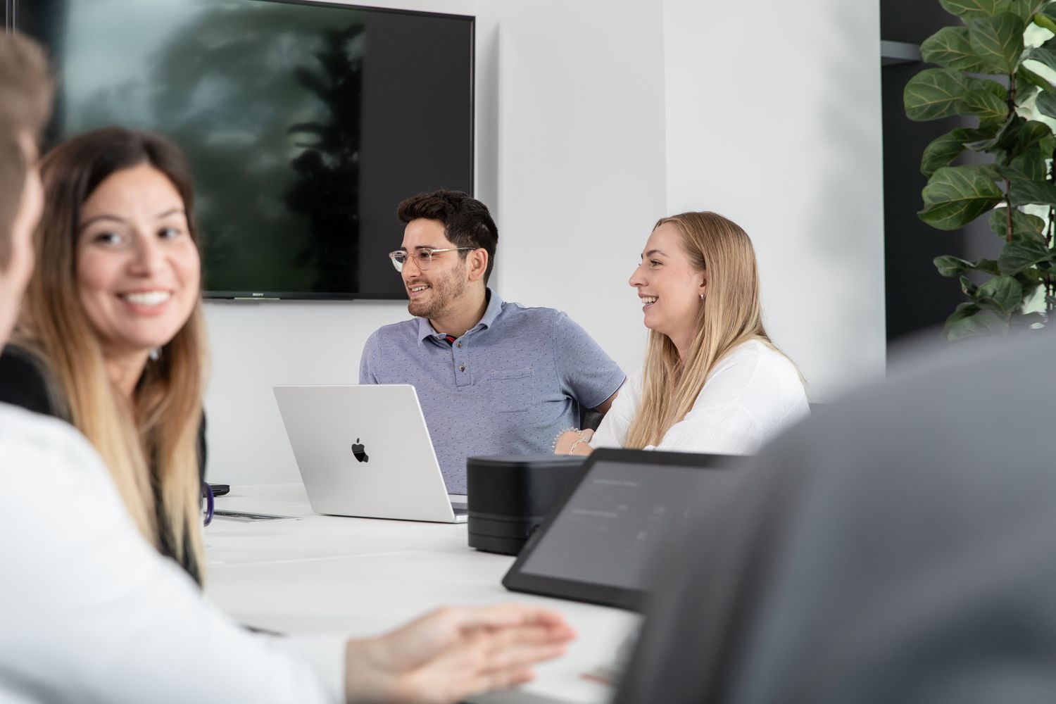 Mehrere Mitarbeiter und Mitarbeiterinnen sitzen im Meetingraum und führen Gespräche, im Mittelpunkt ist ein Apple MacBook zu sehen.